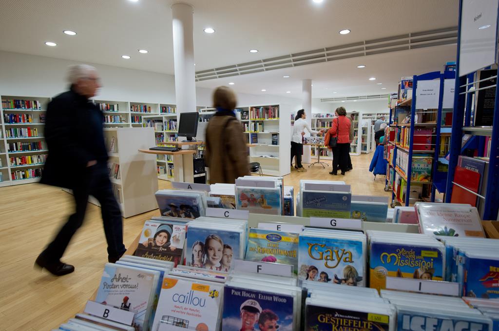 Bücher und DVD-Regale in der Bücherei. Kunden gehen den Mittelgang entlang und werfen einen Blick auf die Regale und einen Infotisch